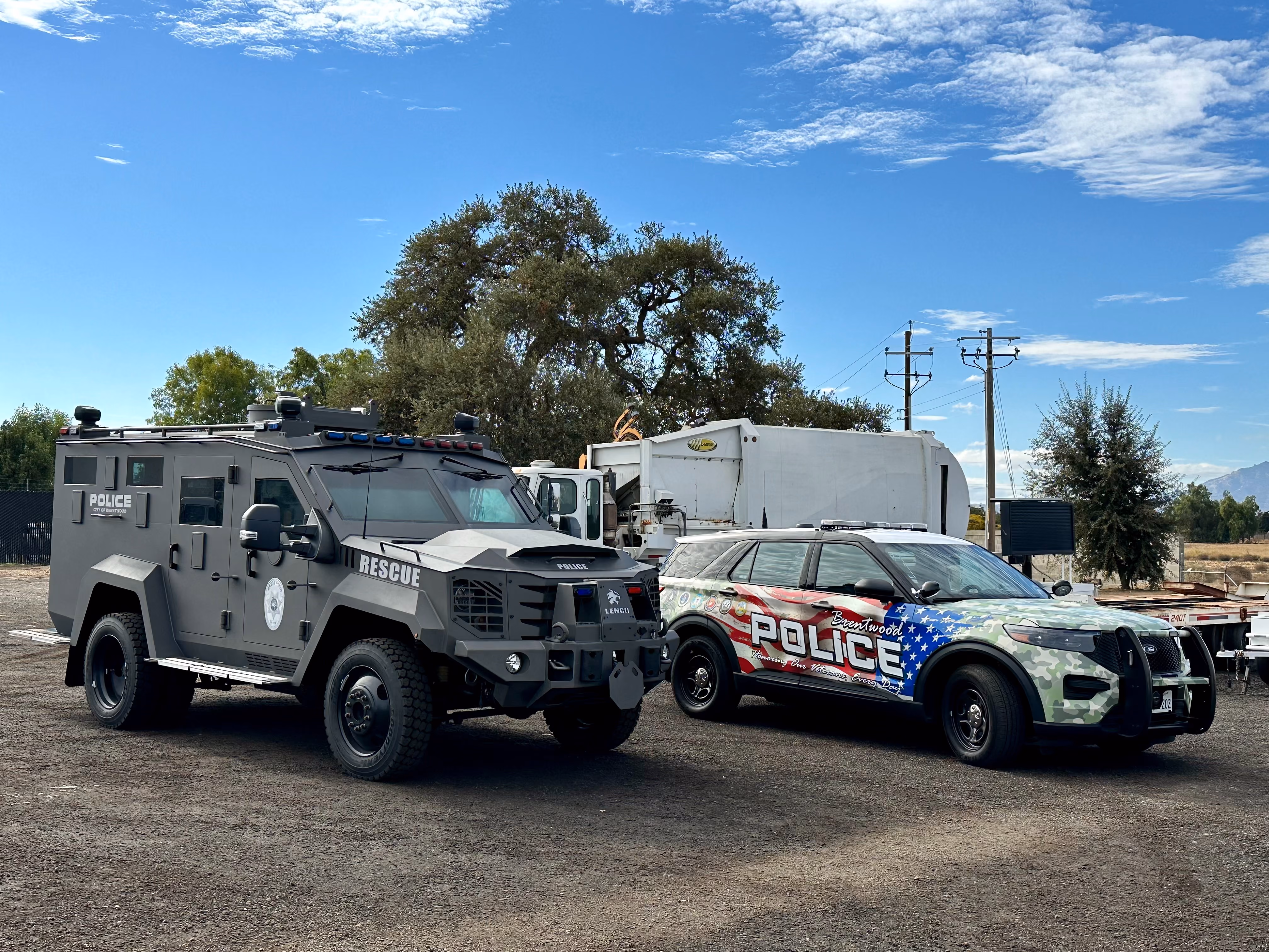 Two police vehicles parked side by side on a dirt ground under a partly cloudy sky. On the left is a matte black armored rescue vehicle with 'POLICE CITY OF BRENTWOOD' and 'RESCUE' markings. On the right is a police SUV with a camouflage and American flag wrap displaying the words 'Brentwood POLICE Honoring Our Veterans Every Day.' Trees, utility poles, and a white truck are in the background.