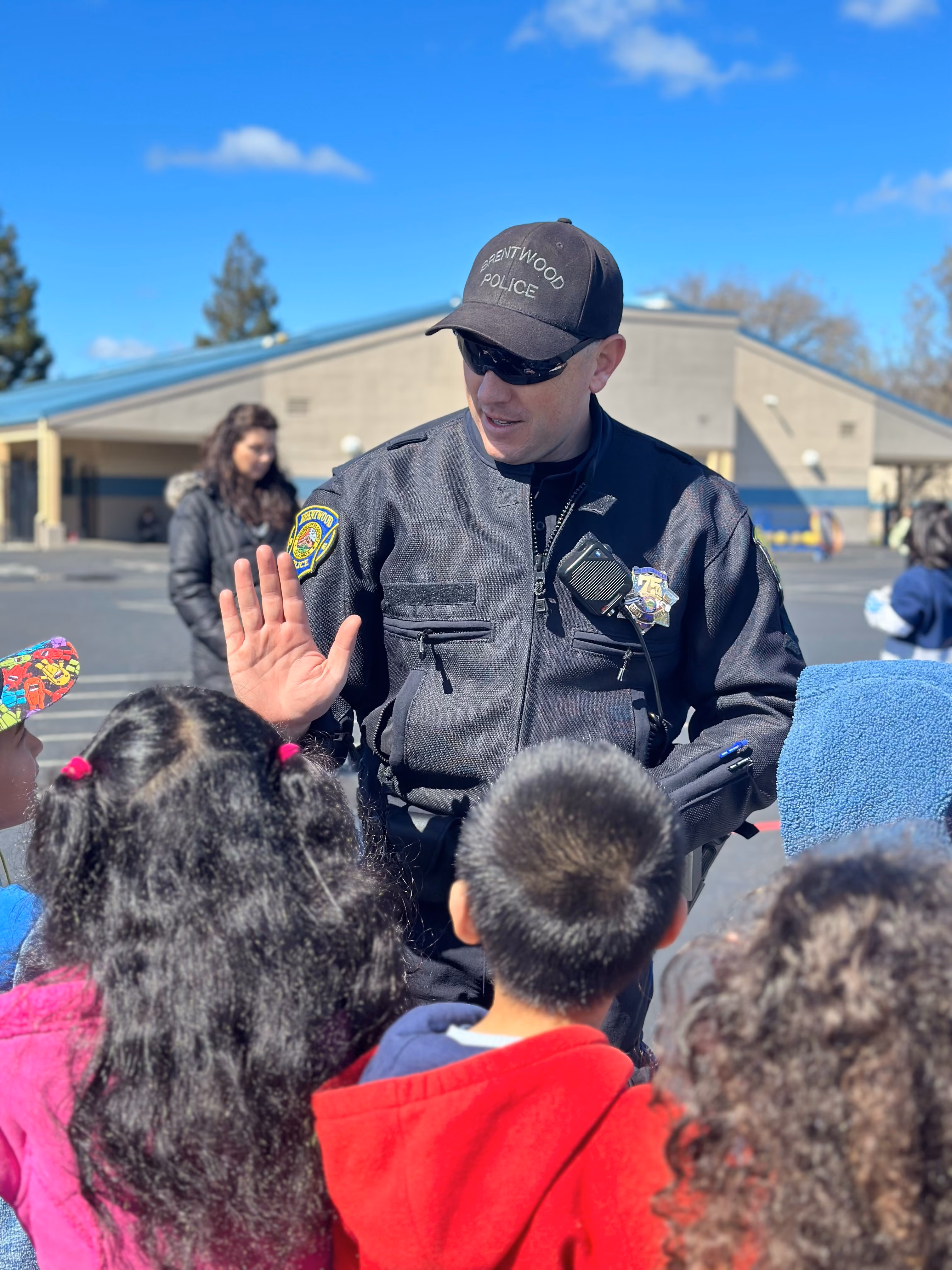 A Brentwood police officer wearing sunglasses and a black uniform is interacting with a group of children outdoors on a sunny day, raising his hand in greeting or to give a high-five.