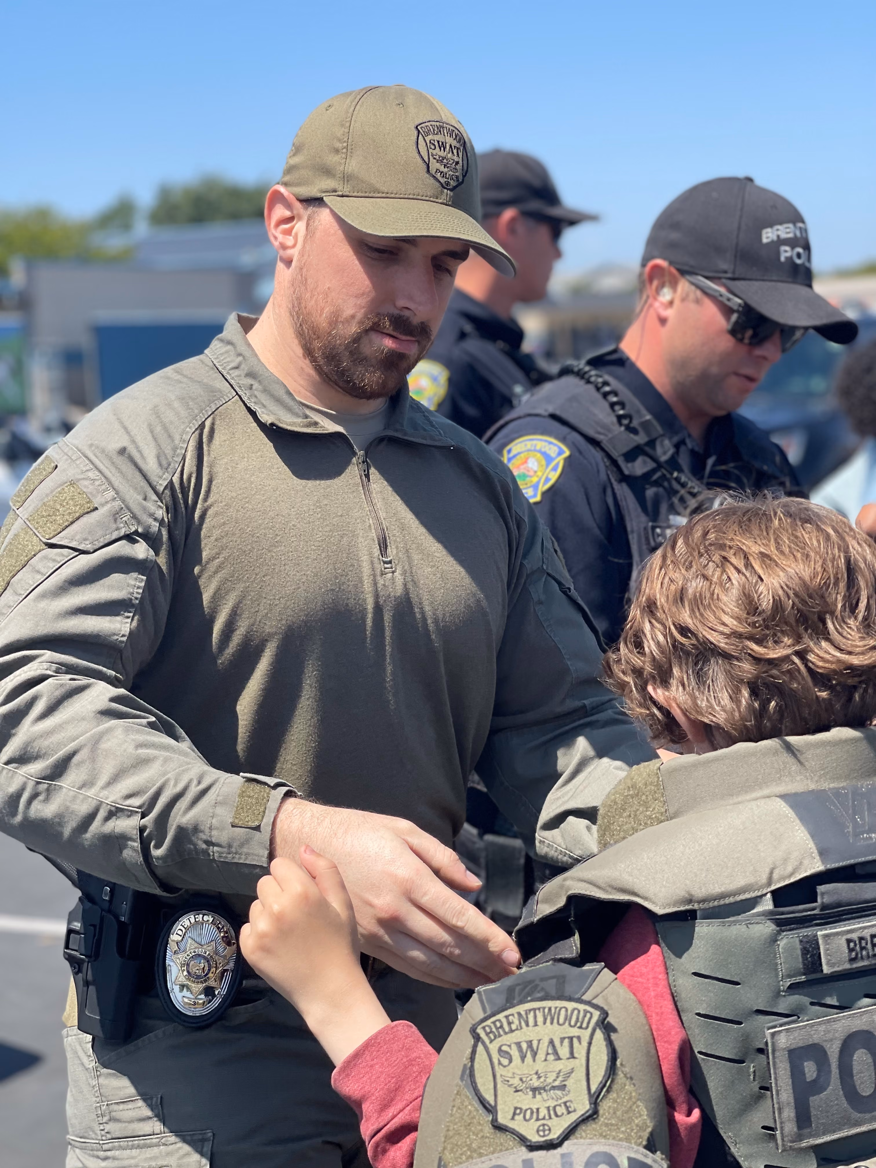 A Brentwood SWAT officer in olive tactical gear and cap helps a child wearing police protective gear outdoors on a sunny day, with two other officers in the background.