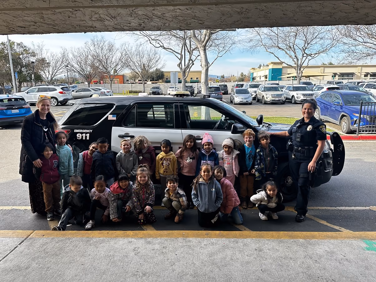 A group of young children, accompanied by a woman and a female police officer, poses in front of a black-and-white police SUV parked in a parking lot with other cars and leafless trees in the background.