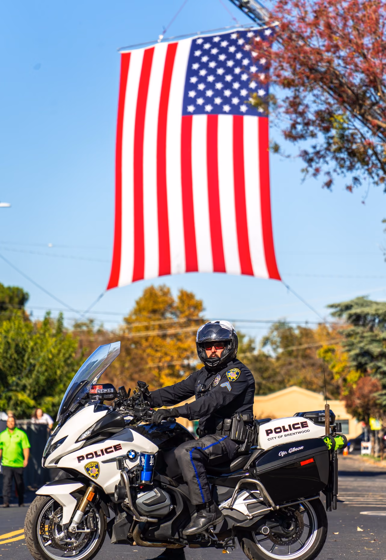A police officer wearing a helmet and sunglasses is sitting on a white police motorcycle with 'Police City of Brentwood' written on it, positioned under a large American flag hanging above the street, with trees and buildings in the background.