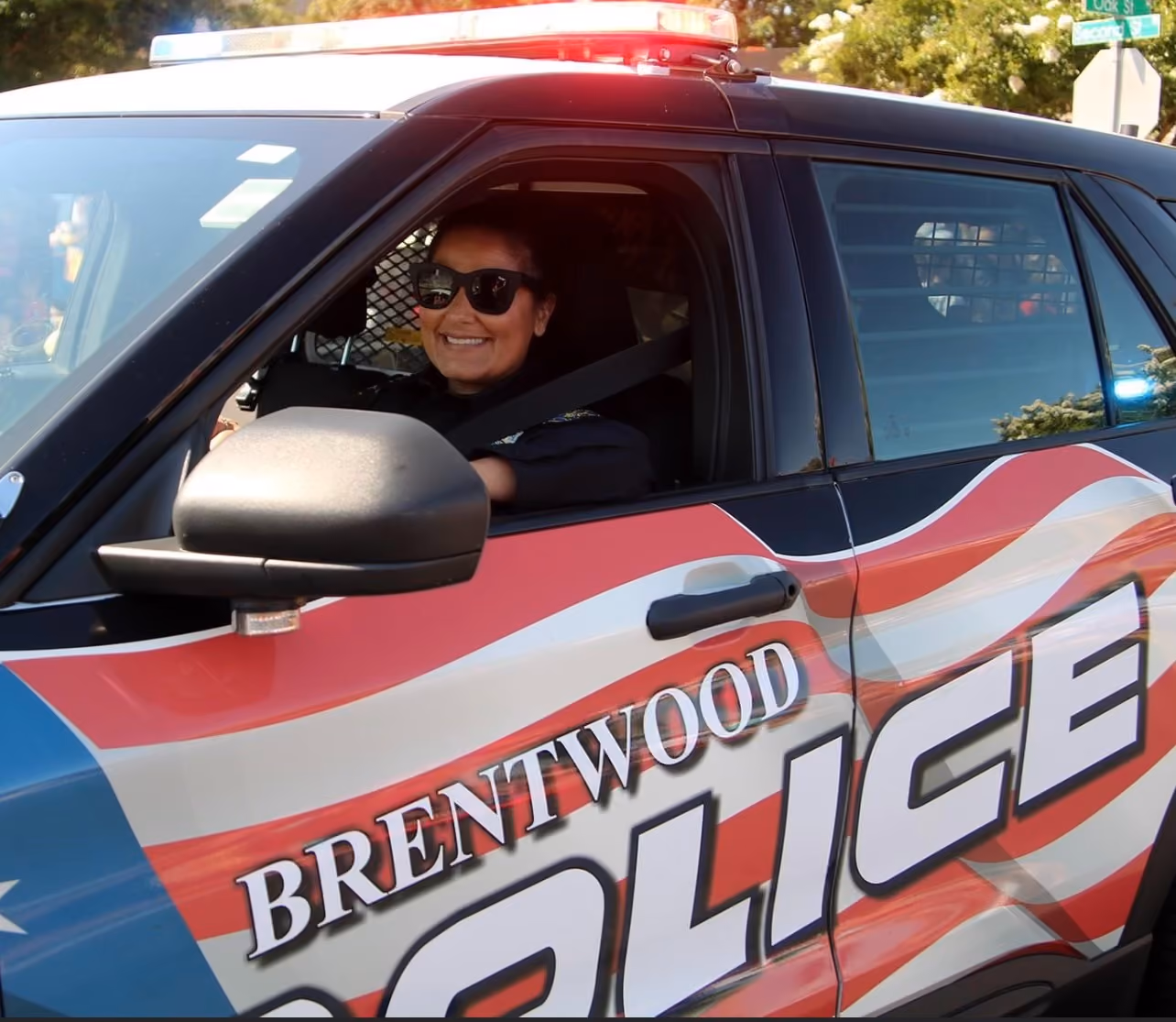 A smiling police officer wearing sunglasses seated inside a Brentwood police car with red, white, and blue markings and a light bar on top.