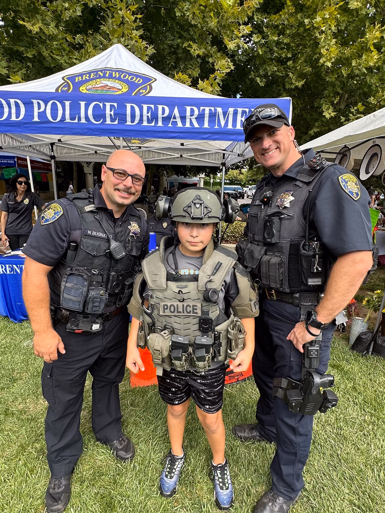 Two police officers in uniform standing on grass with a young boy wearing a police tactical vest and helmet, posing in front of a Brentwood Police Department tent.