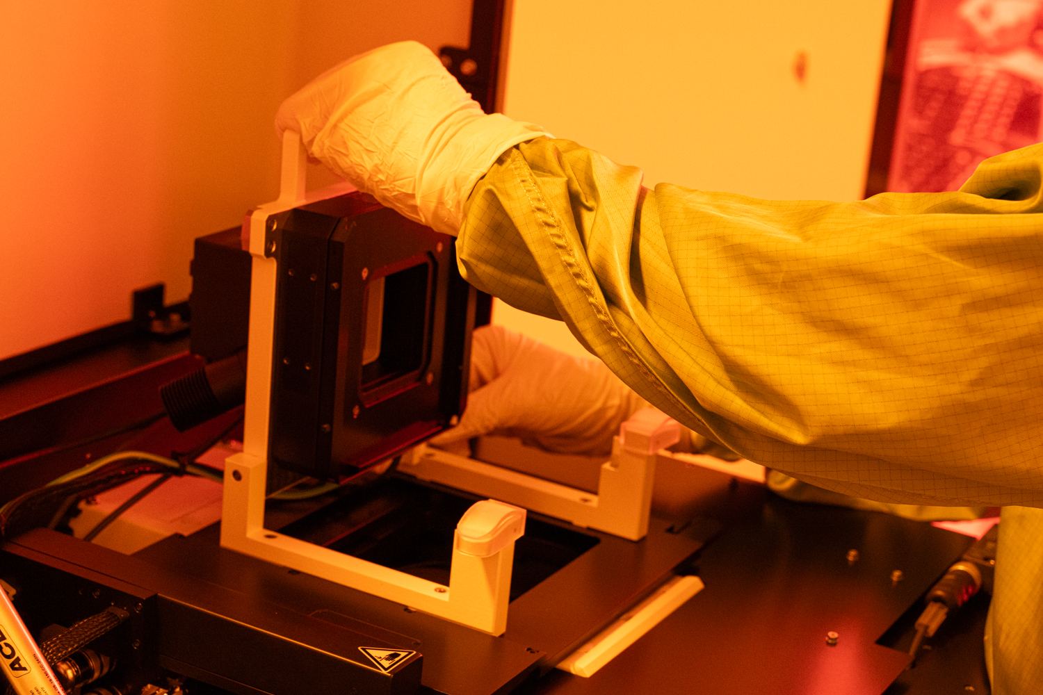 A gloved operator loading a sample holder into a microfabrication tool under the amber light of the clean room.