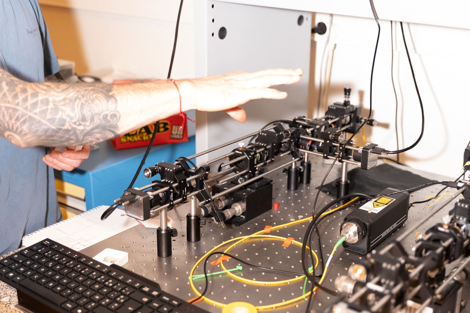 An engineer reaching over the optical characterization bench, lenses and fibres lined up on the table.