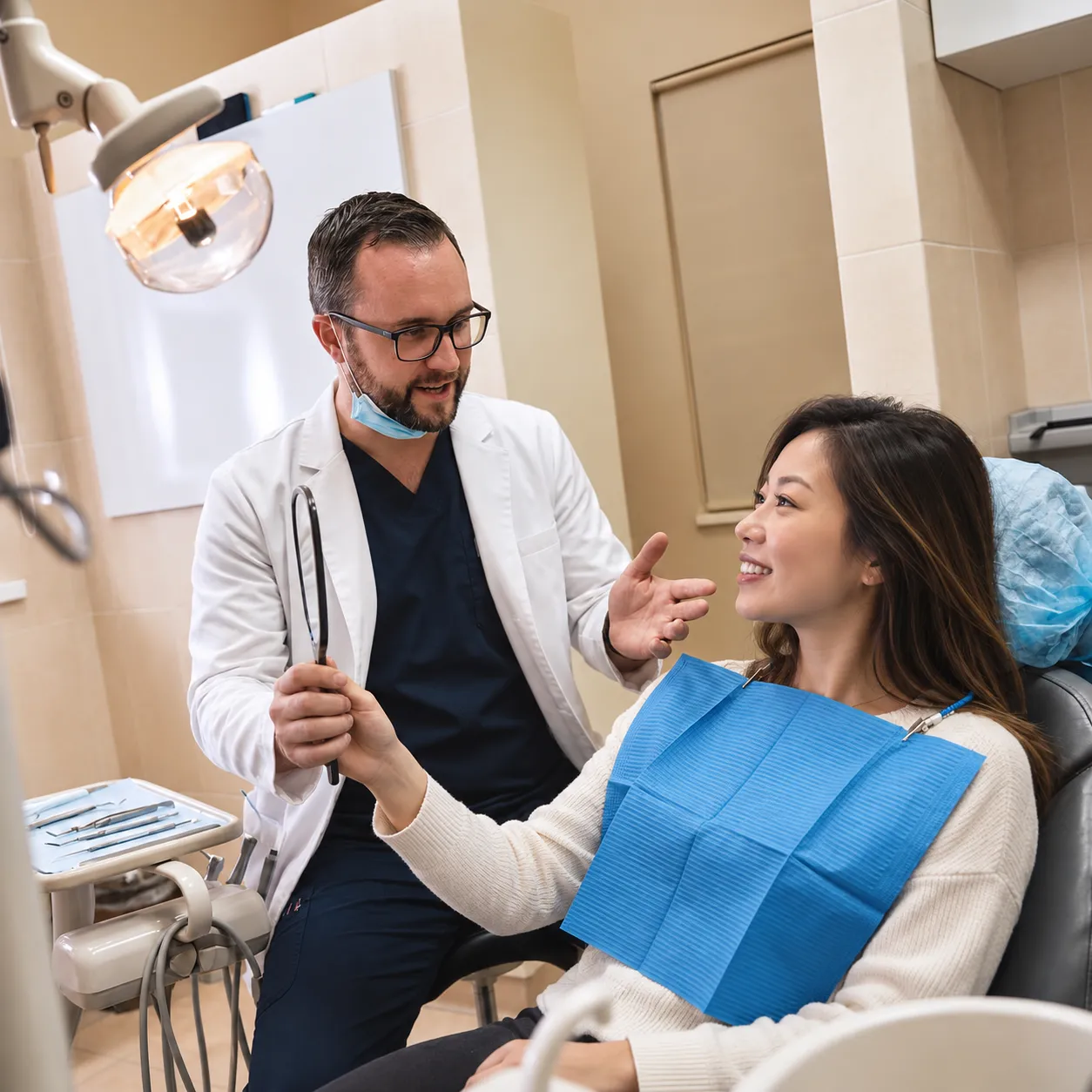 Dentist discussing treatment results with a smiling Asian woman holding a mirror in a modern dental office.