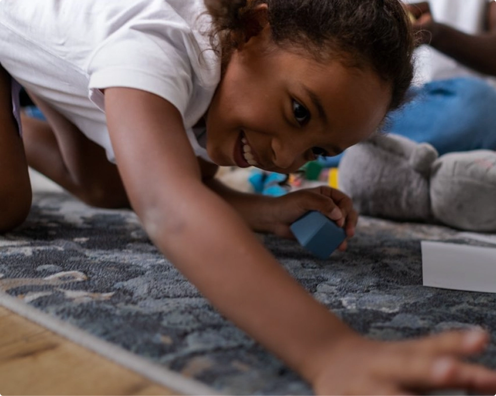 A smiling young child with curly hair playing on a carpet while reaching forward and holding a blue block in one hand.