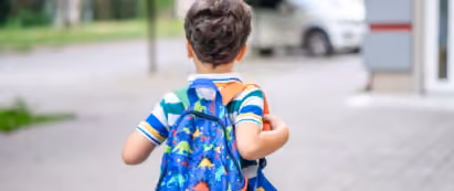Young child wearing a striped shirt and a colorful backpack walking on a sidewalk with blurred background.