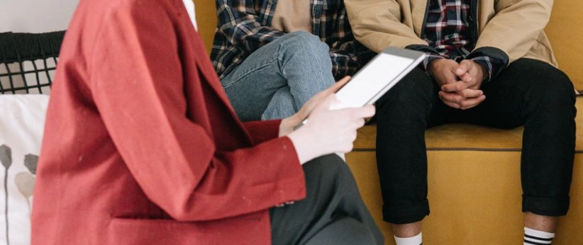Person in a red blazer holding a tablet with two people sitting on a yellow couch in the background.