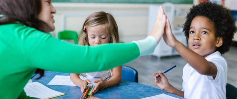 A teacher in a green sweater giving a high-five to a young boy holding a blue pencil at a classroom table, with a young girl sitting beside them holding colored pencils and looking down at her paper.