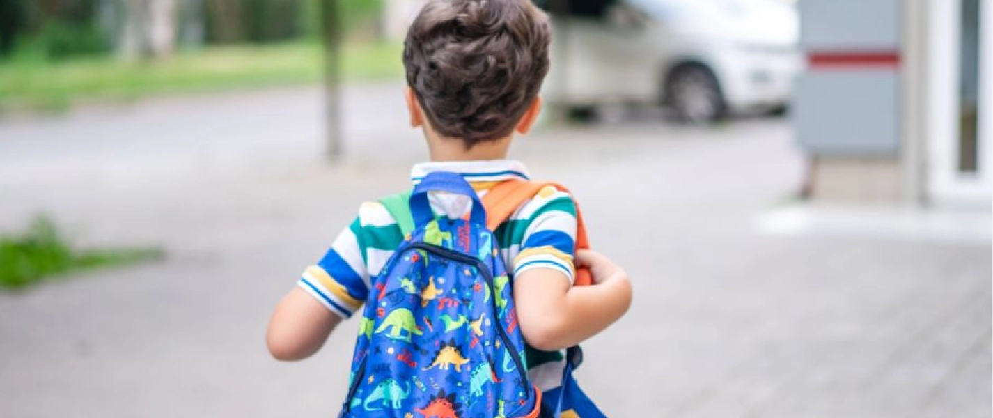 Young child with short brown hair wearing a colorful striped shirt and a blue dinosaur backpack walking outdoors on a paved path.