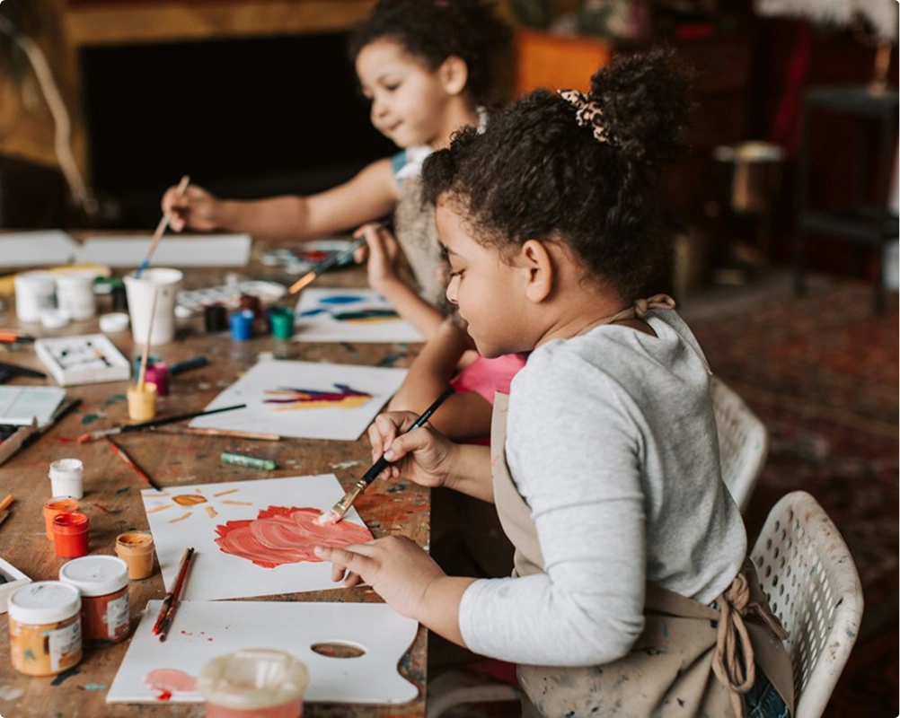 Two young girls with curly hair wearing aprons sit at a wooden table painting with brushes and colorful paints on paper.