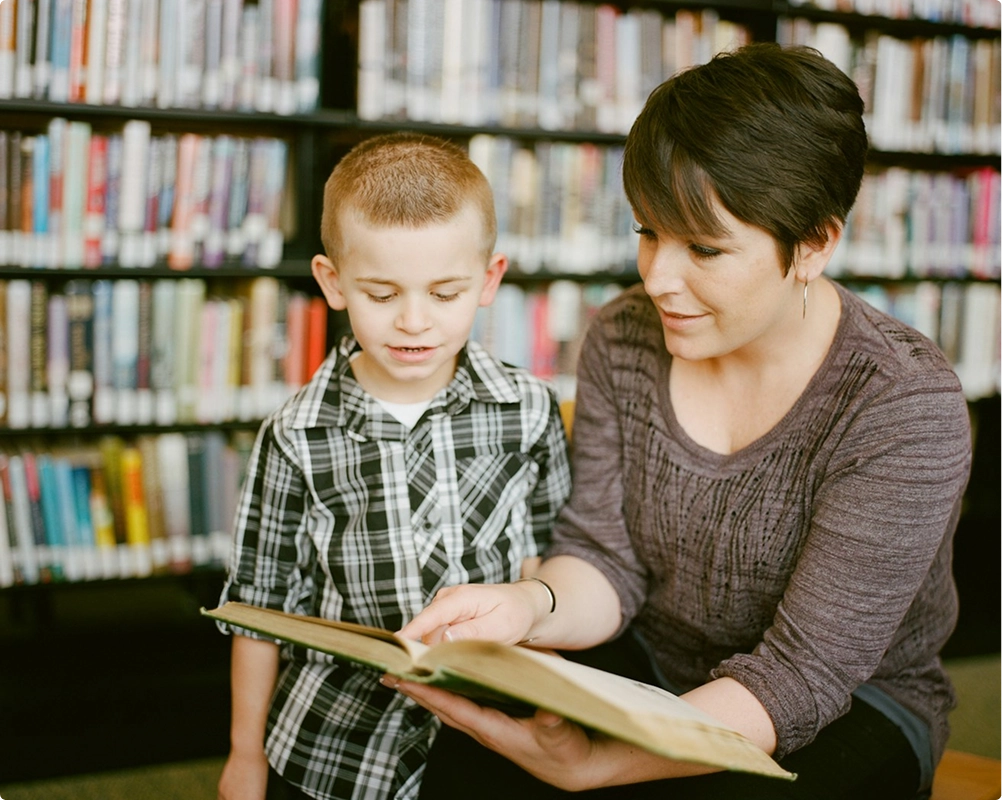 A woman with short dark hair is reading a book to a young boy in a plaid shirt in a library, with bookshelves in the background.