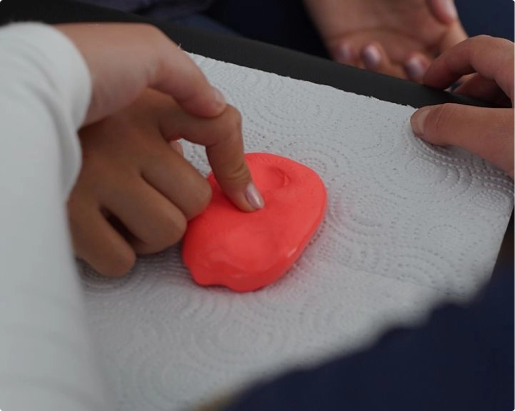 Two hands pressing fingers into a flattened ball of bright red clay on a white paper towel.