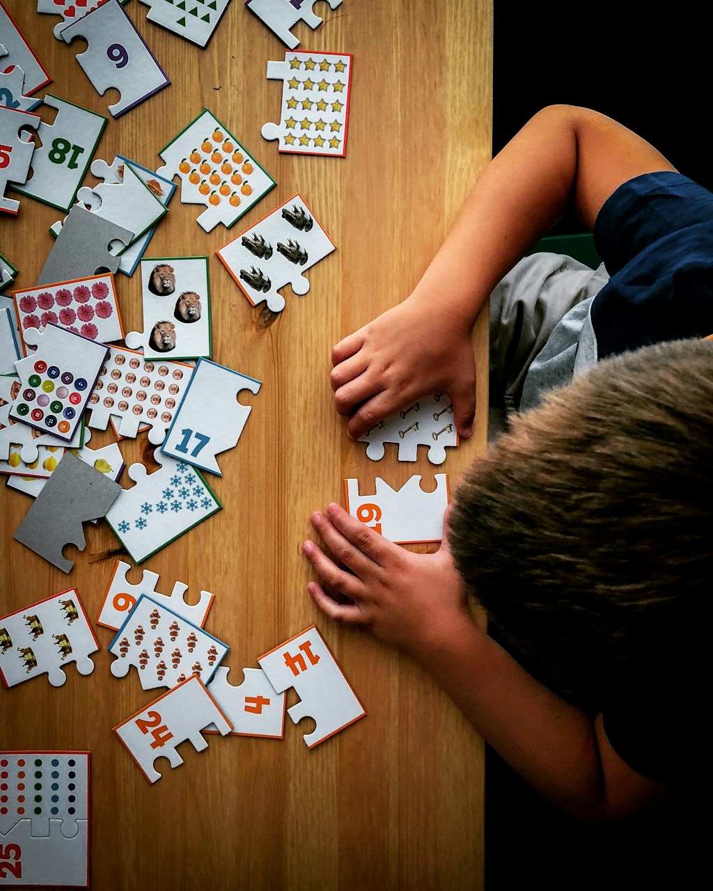 Top-down view of a child assembling a wooden puzzle on a table with pieces featuring numbers and images, including lions, stars, and buttons.