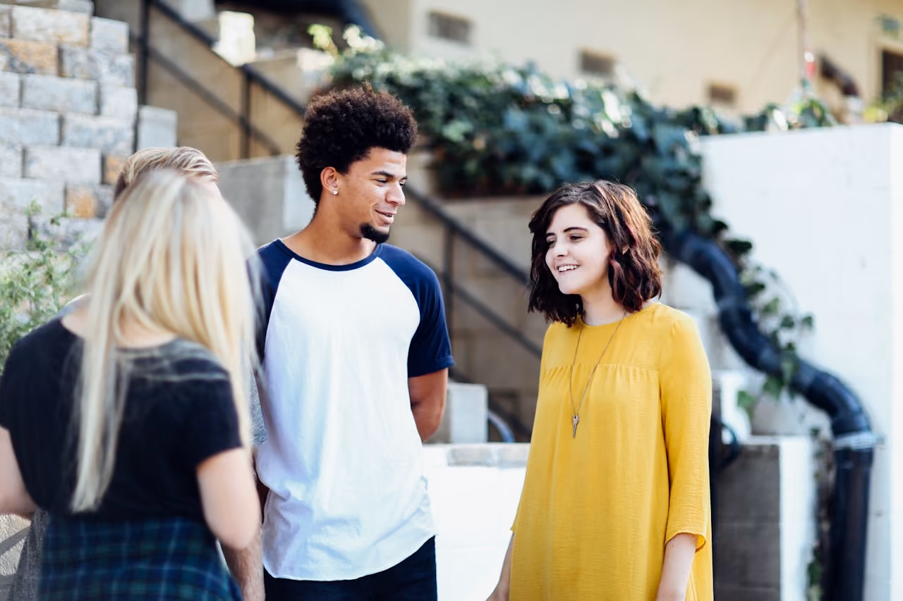Group of young adults casually talking outside near stairs and greenery; one young woman in a yellow dress smiles while a young man in a white and navy shirt looks at her.