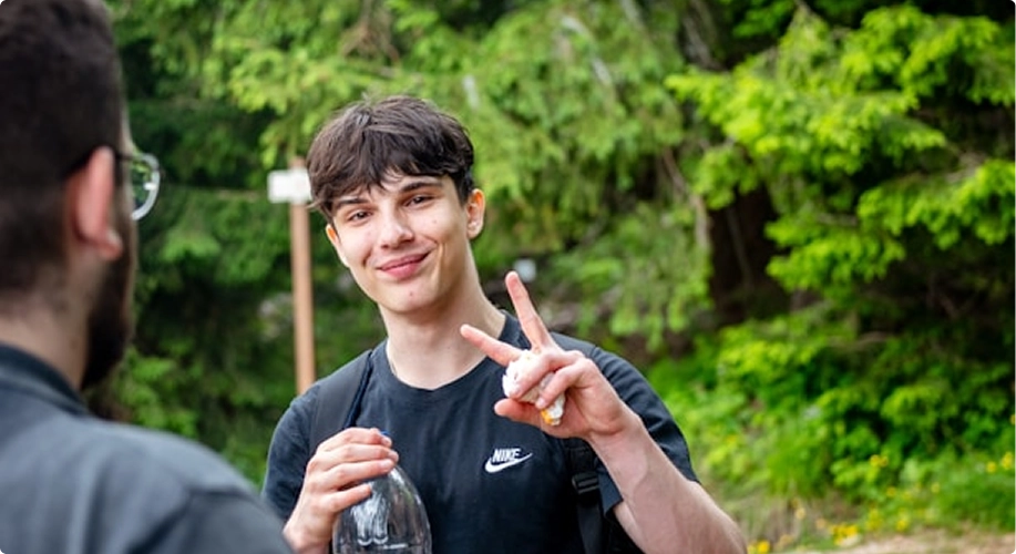 Young man wearing a black Nike t-shirt and backpack outdoors, smiling and making a peace sign with his right hand, holding a clear plastic bottle in his left hand, with another person partially visible in the foreground, green trees in the background.