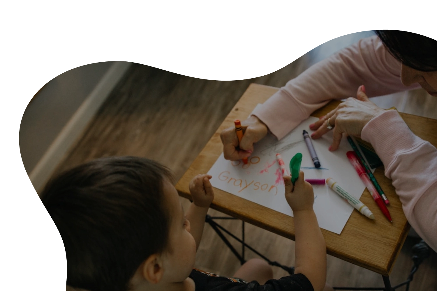 A child and an adult sitting at a small wooden table with crayons, drawing and coloring on a piece of paper with the name Grayson written in orange crayon.