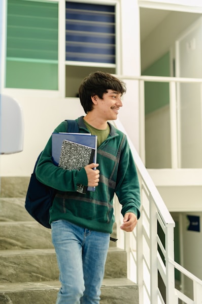 A young male student wearing a green sweater and blue jeans descends stairs indoors, carrying a backpack and holding notebooks, smiling and looking to his right.