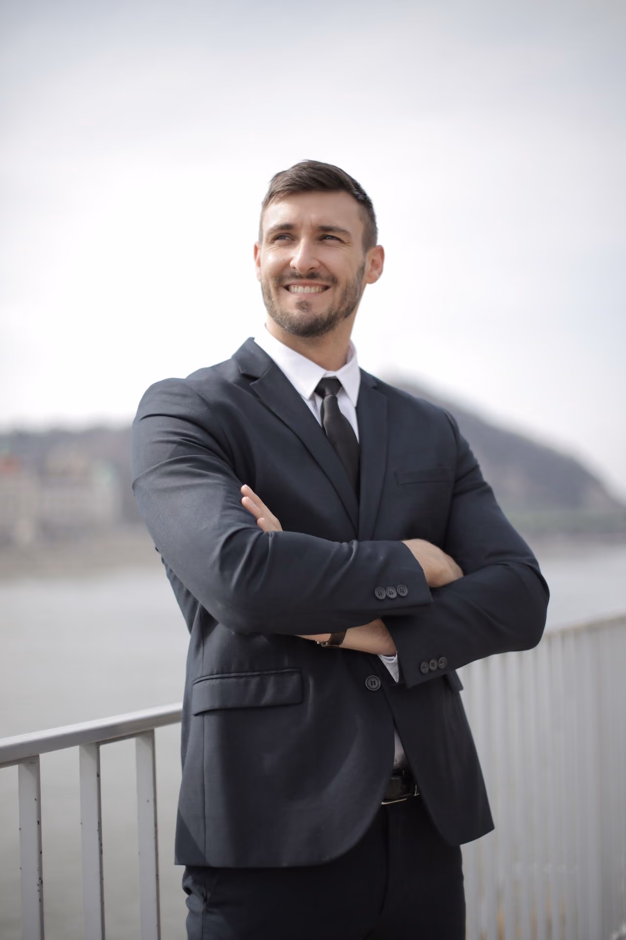 Retrato de un ejecutivo sonriente en traje con los brazos cruzados al aire libre. Representa la confianza, el liderazgo y la certeza en la toma de decisiones comerciales.
