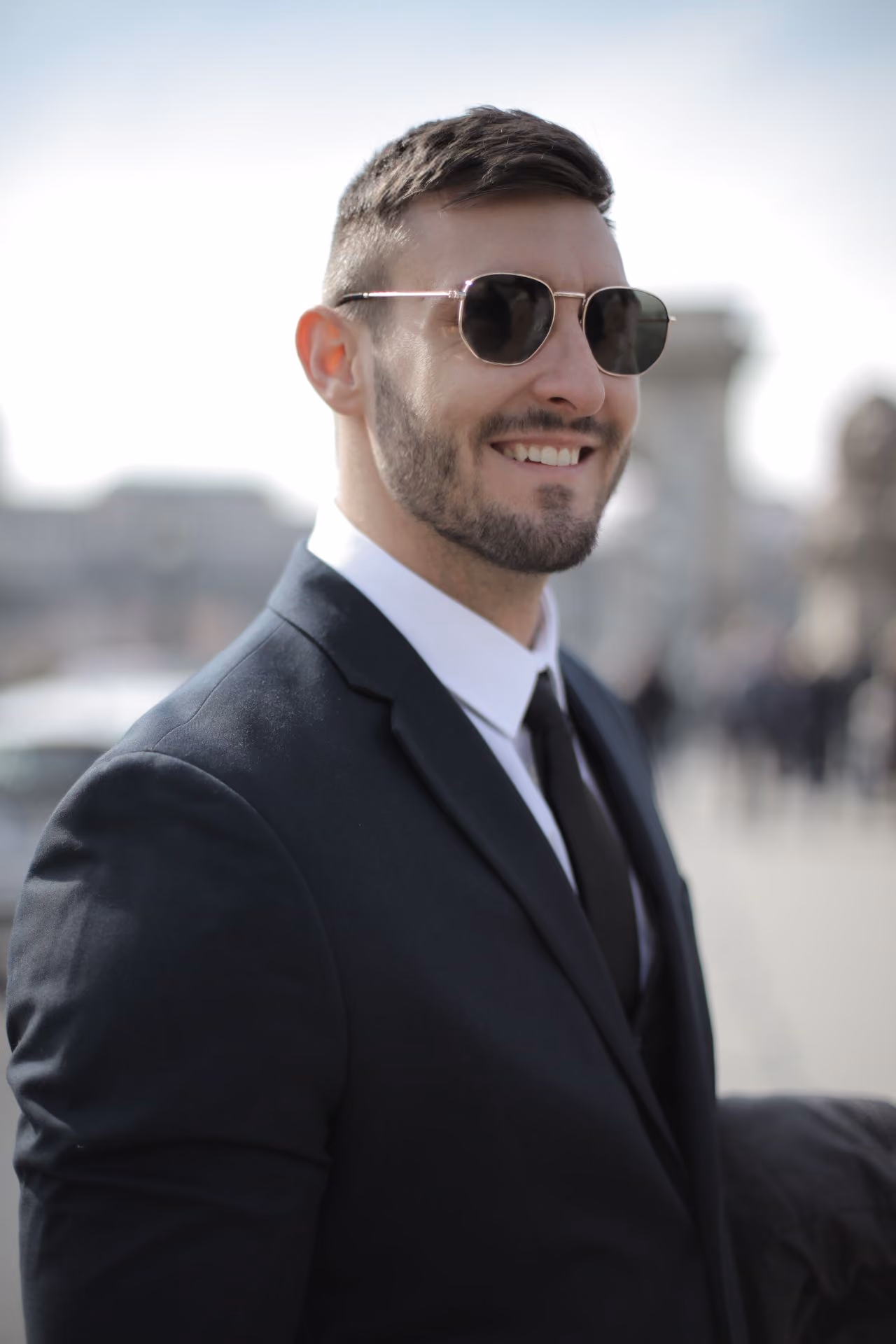 Hombre joven con barba y gafas de sol negras, vestido con traje oscuro, camisa blanca y corbata negra, sonriendo en un entorno urbano desenfocado.