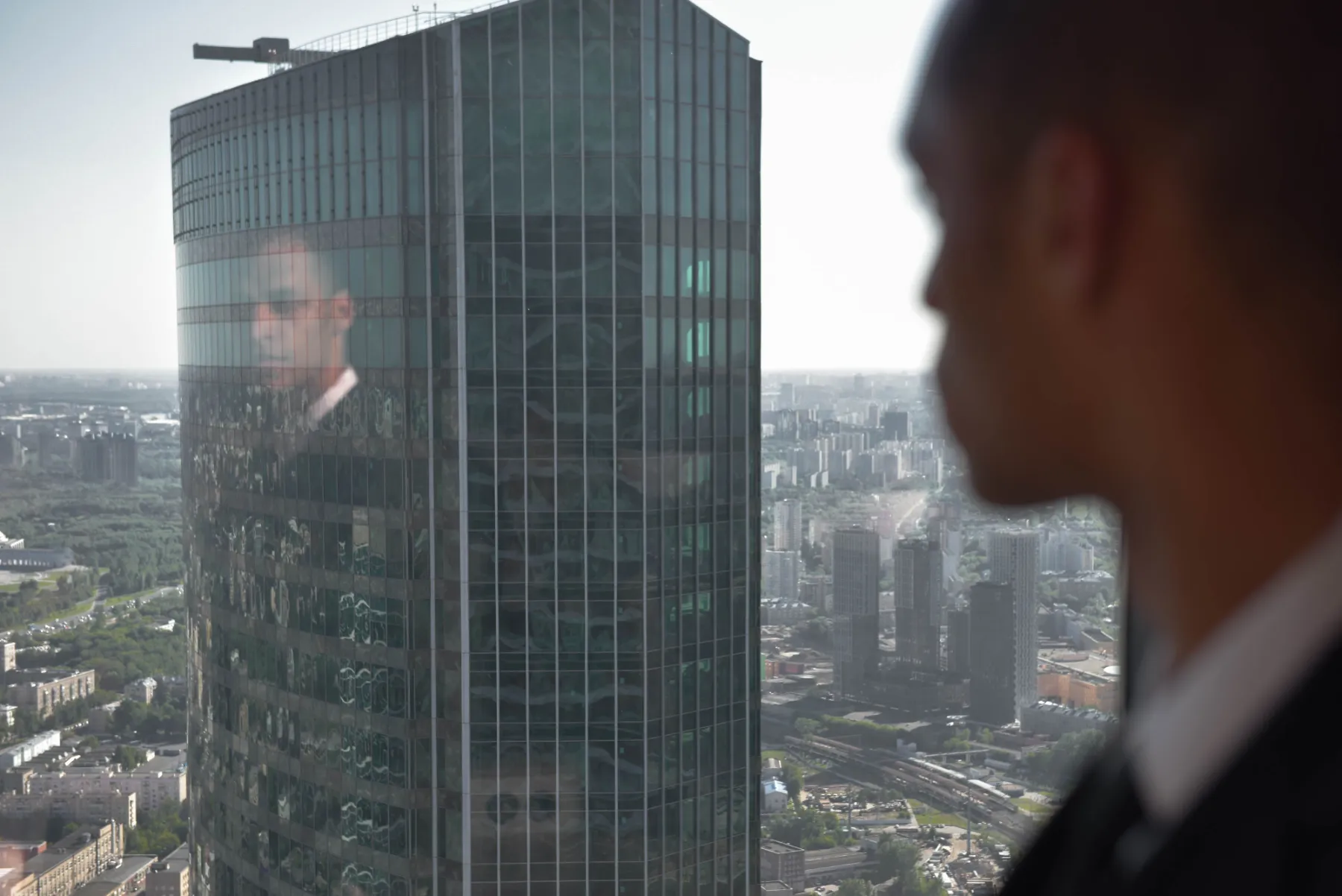 Man in a suit reflected in the glass of a high-rise, overlooking the city.