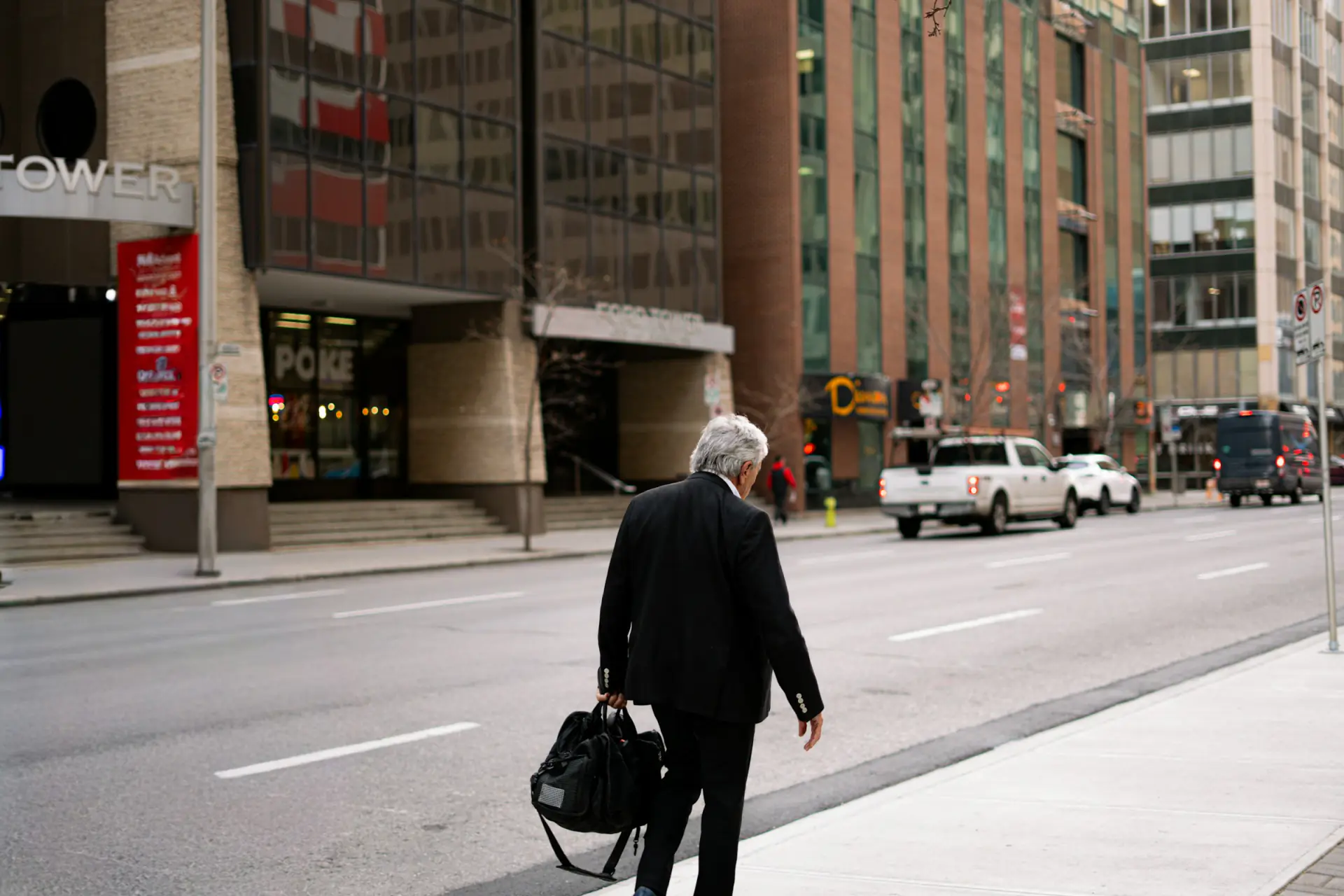 Grey-haired man in a dark suit carrying a bag along a city street lined with office towers.
