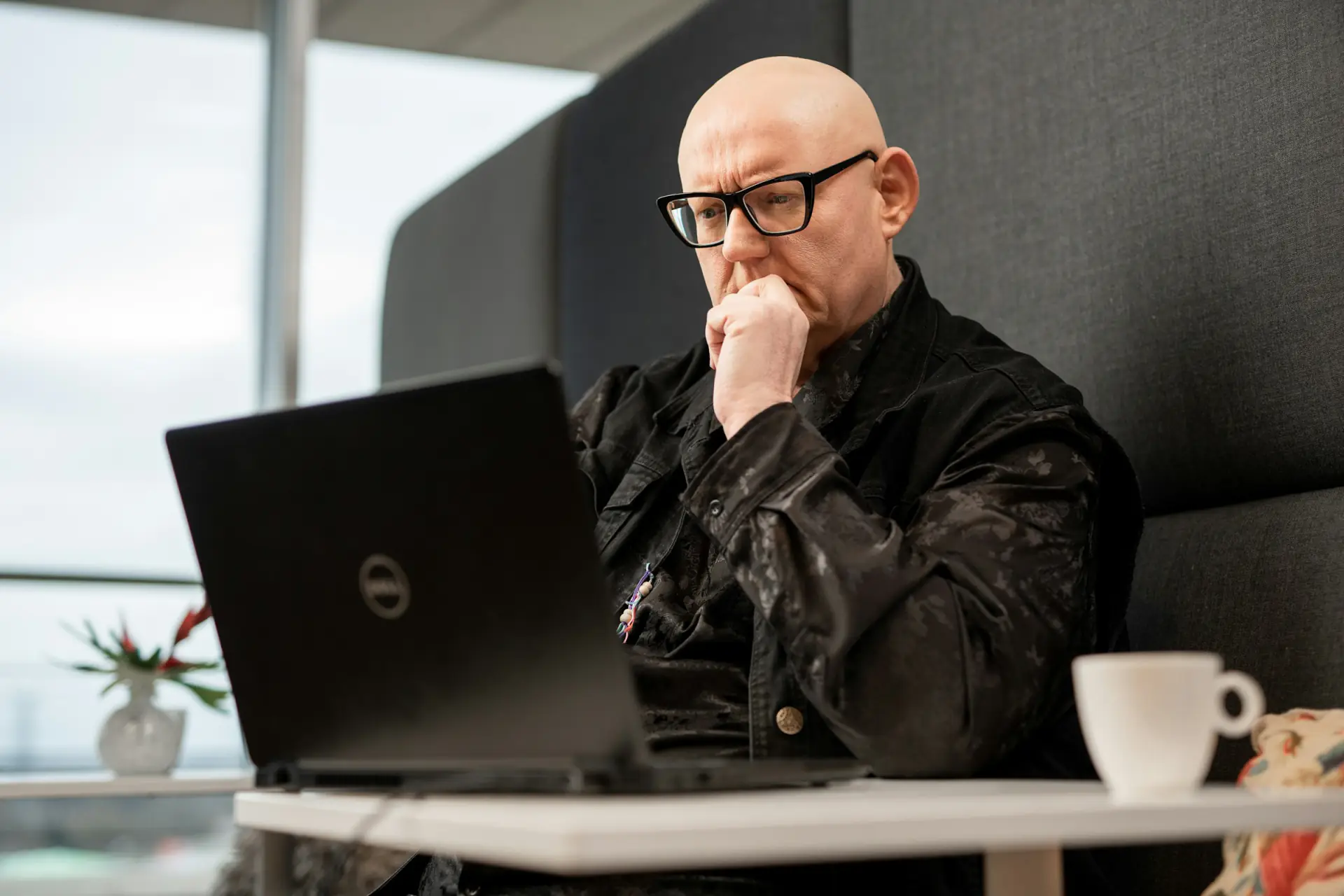 Bald man in glasses and a dark jacket, chin in hand, thinking while working at a laptop by a window.