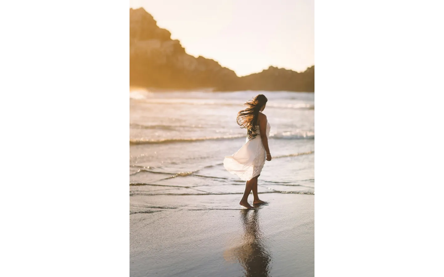 Woman in a white dress walking along the shoreline at sunset