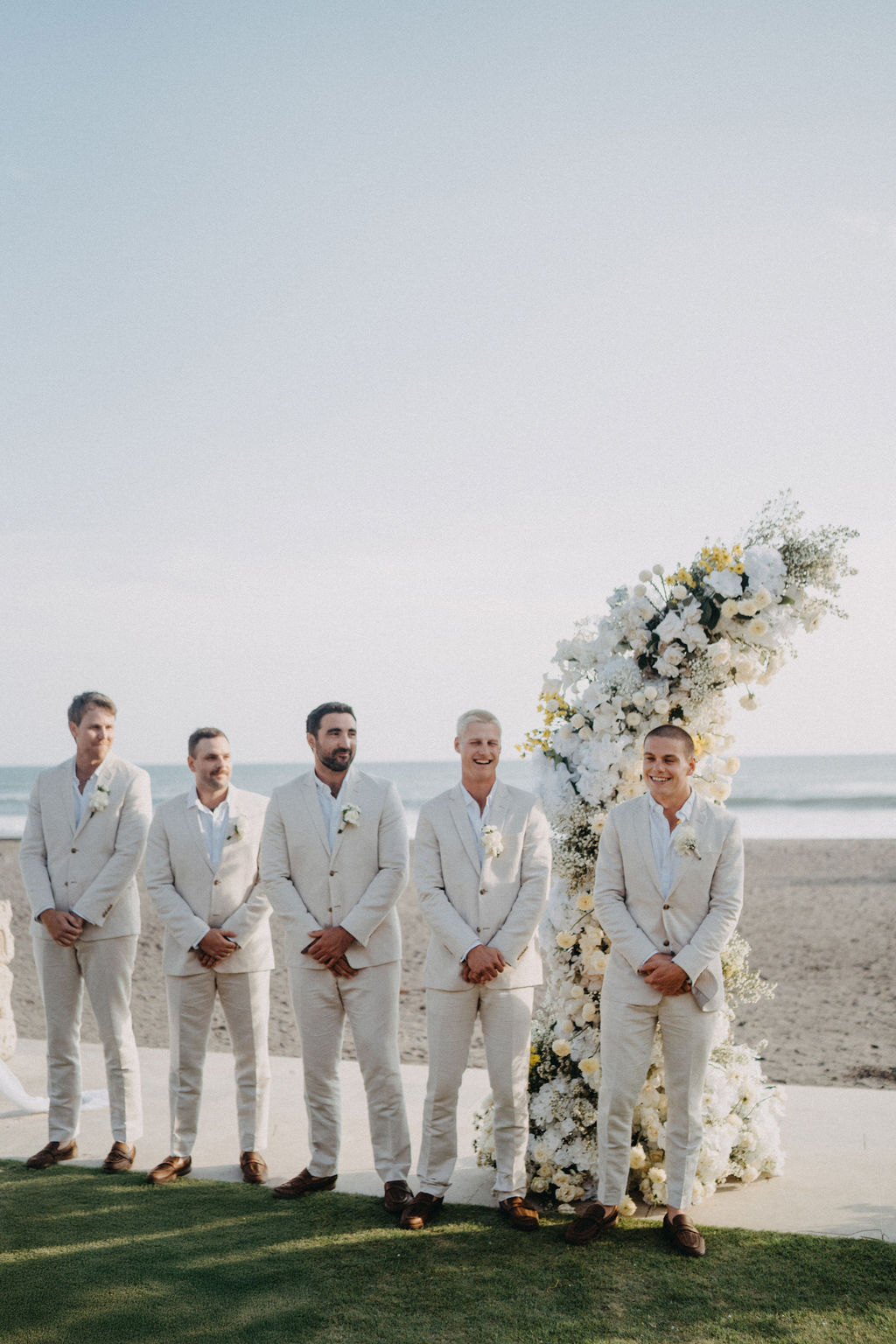 Groomsmen standing beside a white floral wedding arch on the beach