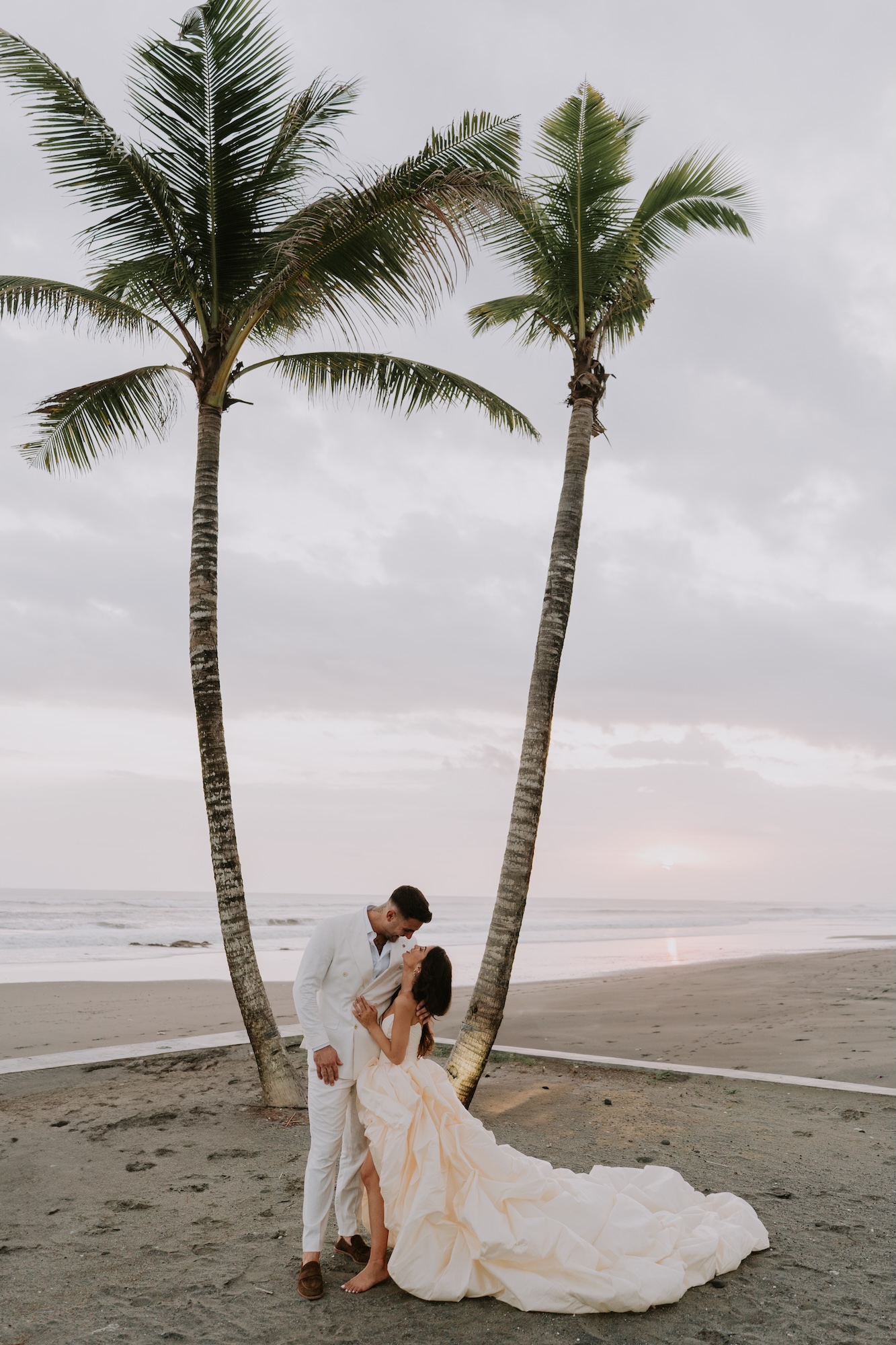 Bride and groom sharing a kiss beneath palm trees on the beach at sunset