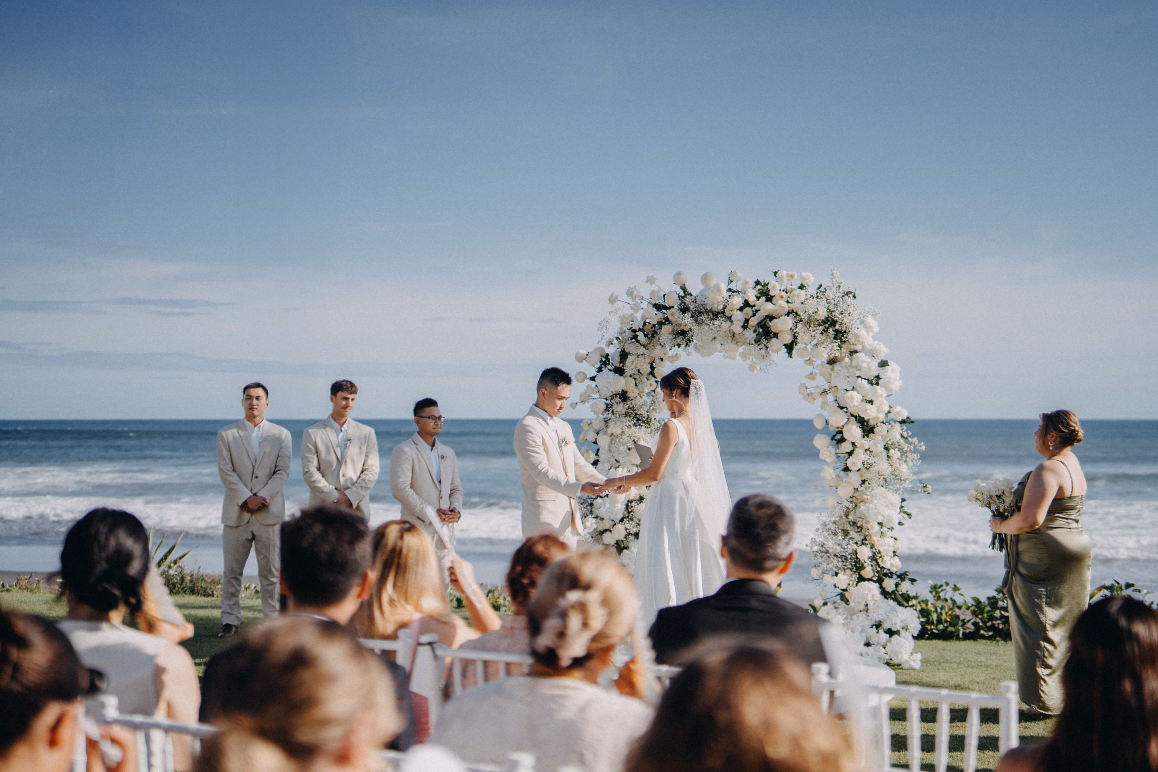 Bride and groom exchanging vows under a floral arch at an oceanfront ceremony
