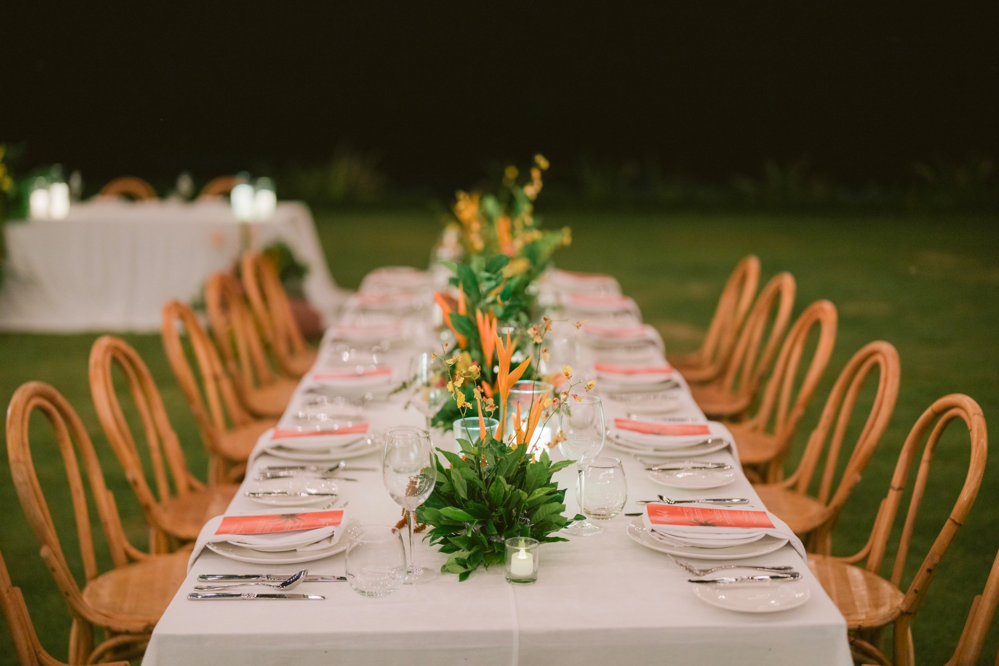 Long reception table with wooden chairs and tropical floral centerpieces in the evening