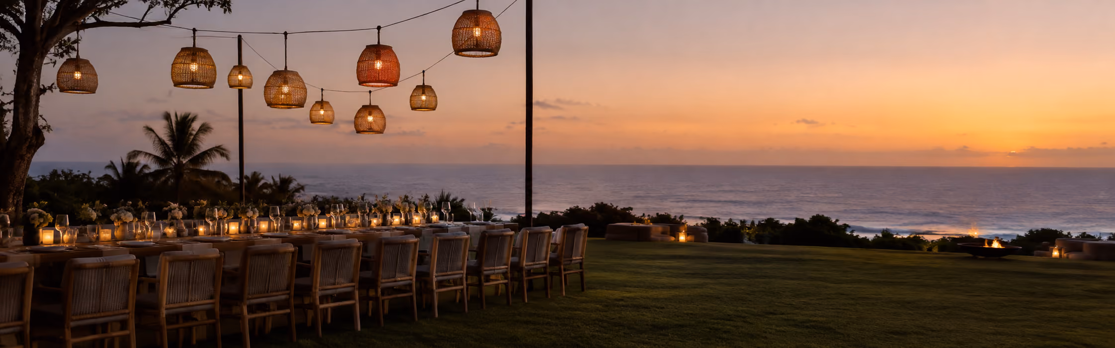 Long dining table strung with hanging lanterns set up on the lawn at sunset