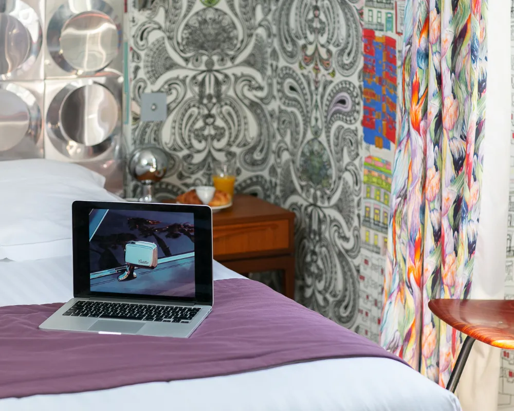 Ordinateur portable posé sur le lit de la Suite Junior de l'Hôtel Crayon à Paris 1er, papier peint baroque et rideaux colorés