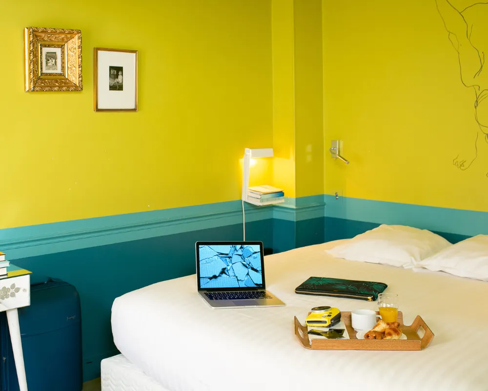 Chambre double jaune et bleu canard de l'Hôtel Crayon à Paris 1er avec ordinateur et petit-déjeuner au lit