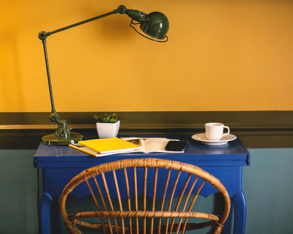 Bureau bleu, lampe vintage et chaise en rotin dans une chambre jaune de l'Hôtel Crayon, Paris 1er