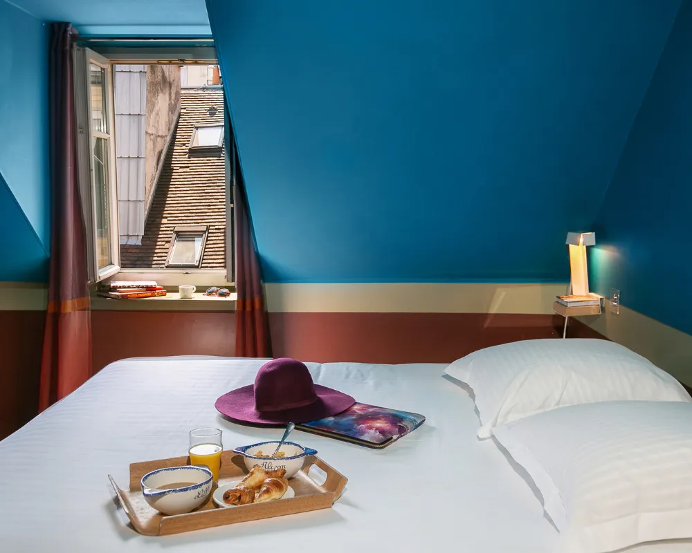 Chambre mansardée bleue de l'Hôtel Crayon avec vue sur les toits de Paris 1er et plateau petit-déjeuner