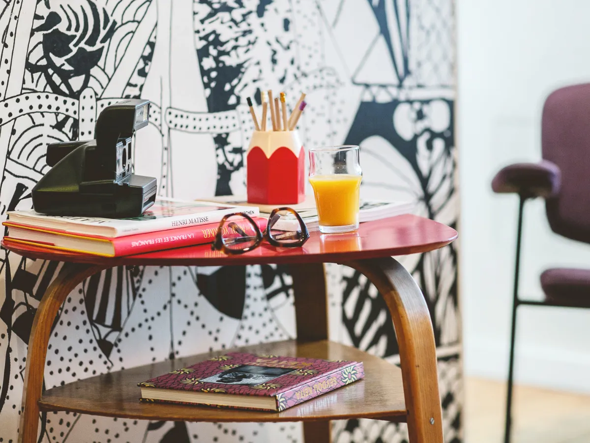 Vintage pedestal table with a camera, pencils, and magazines in front of graphic wallpaper at Hôtel Crayon, Paris