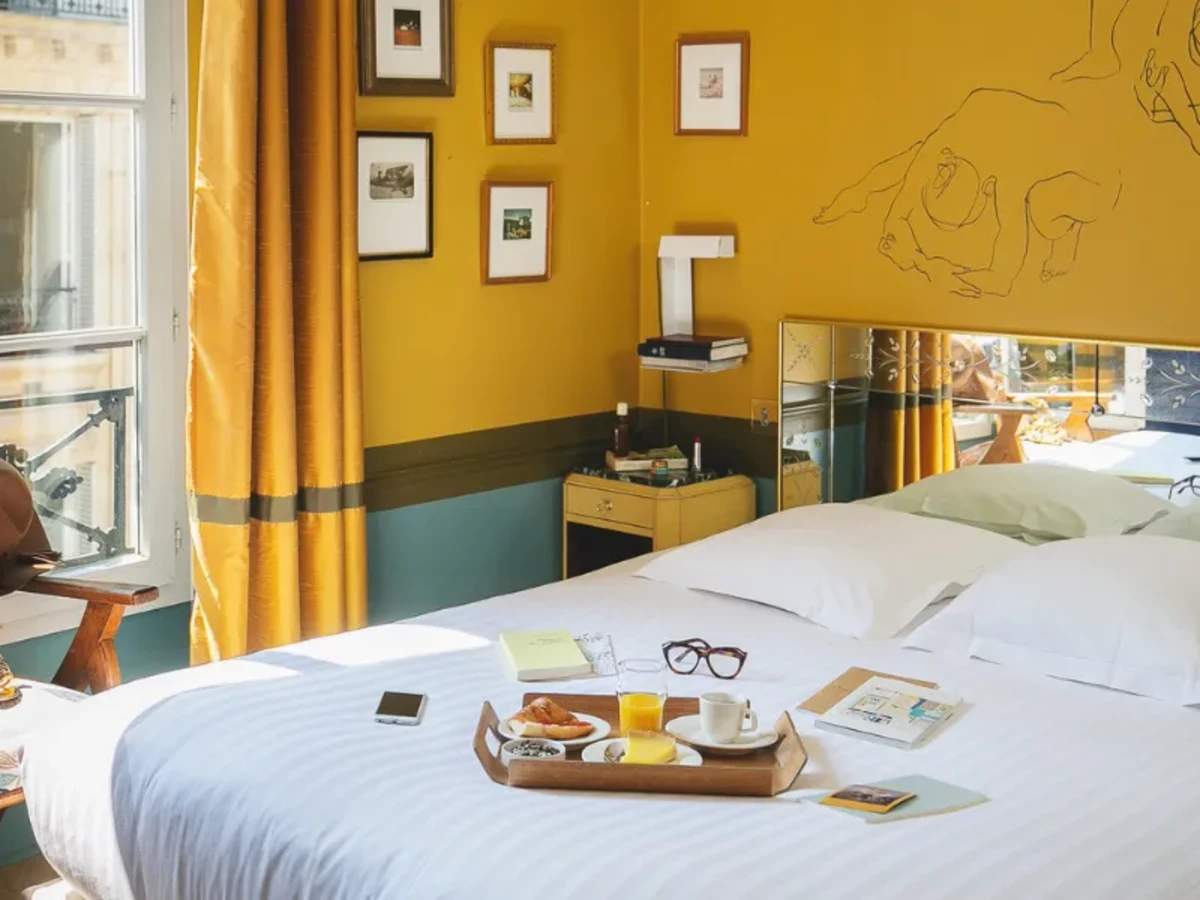 Chambre d'hôtel lumineuse avec un grand lit blanc, plateau de petit-déjeuner avec jus, café, croissant, une paire de lunettes et un téléphone posé dessus, un fauteuil en bois avec un chapeau brun et des chaussures dorées, rideaux jaunes, murs jaunes ornés de cadres et d'un dessin linéaire.