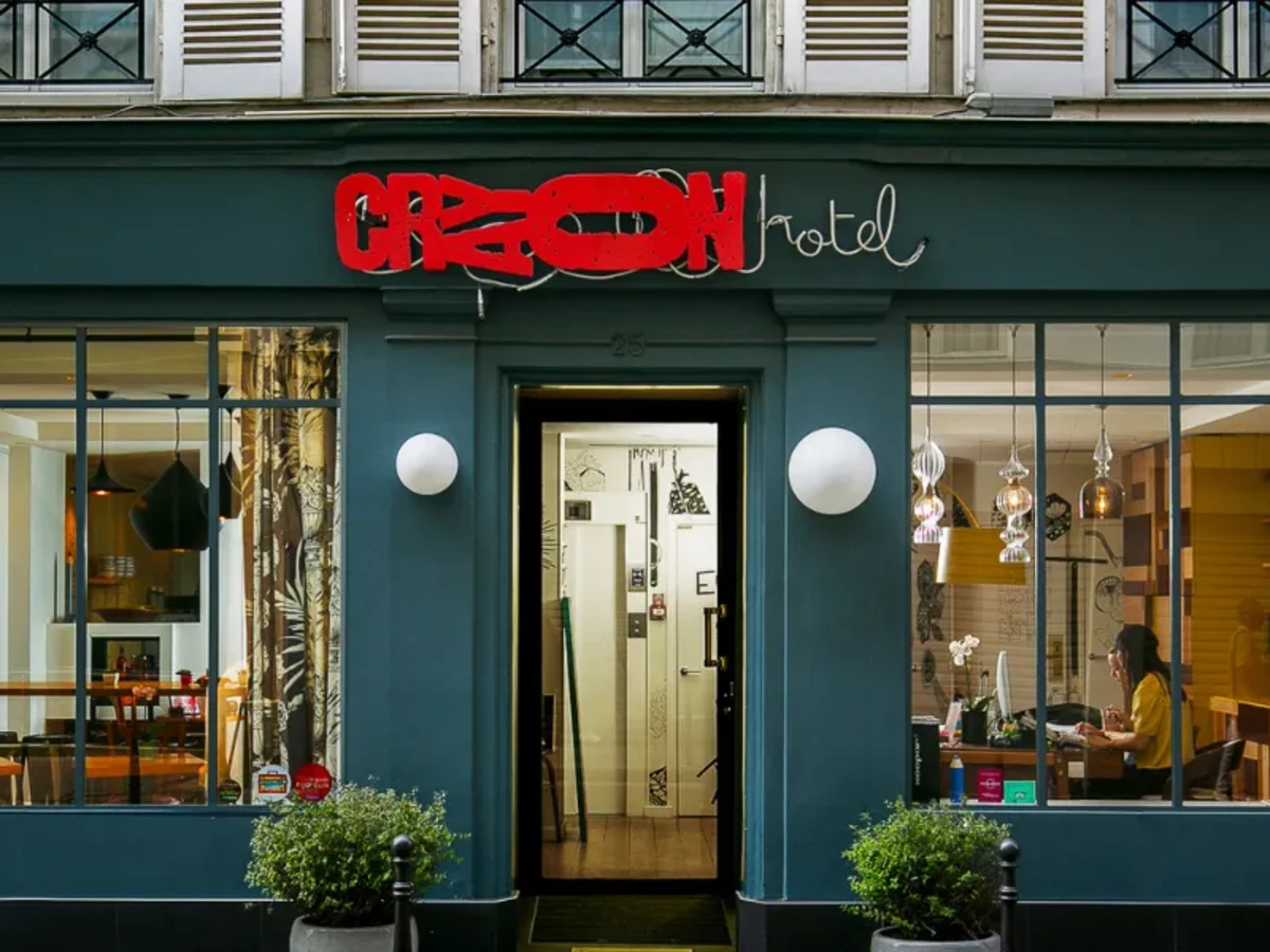Façade bleu canard de l'Hôtel Crayon à Paris 1er, enseigne néon rouge et salons visibles à travers les vitrines