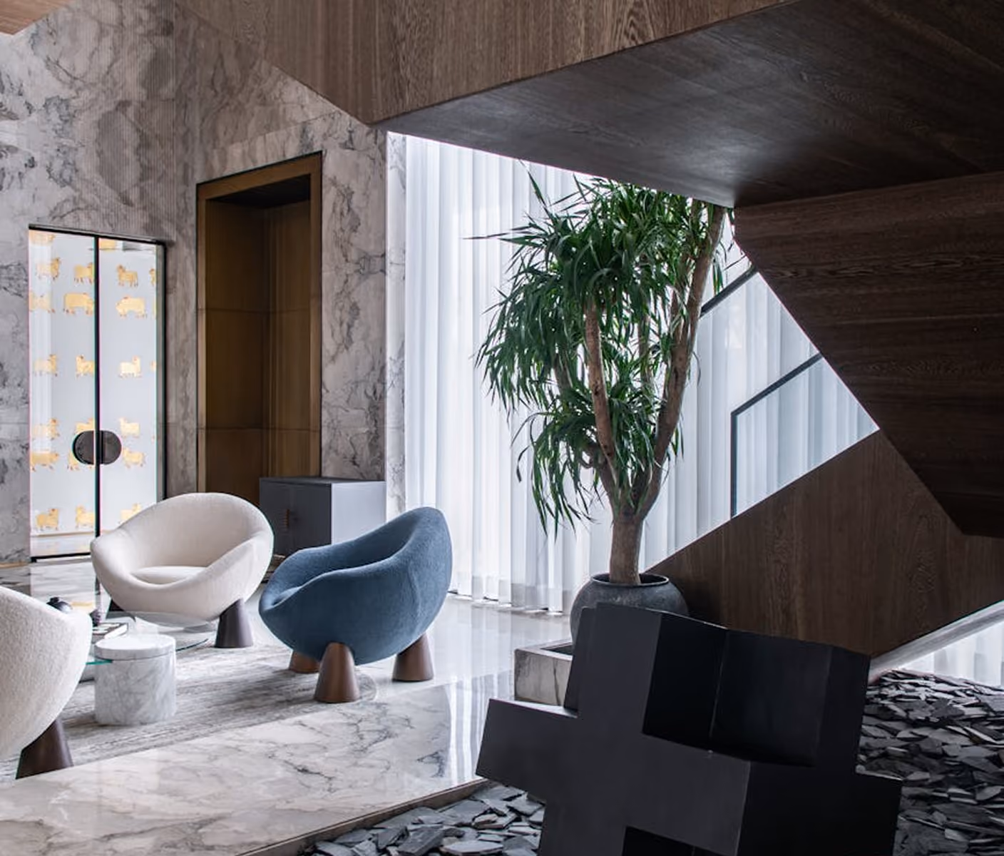Modern living room with curved chairs, marble floor, large plant, and dark wood staircase.