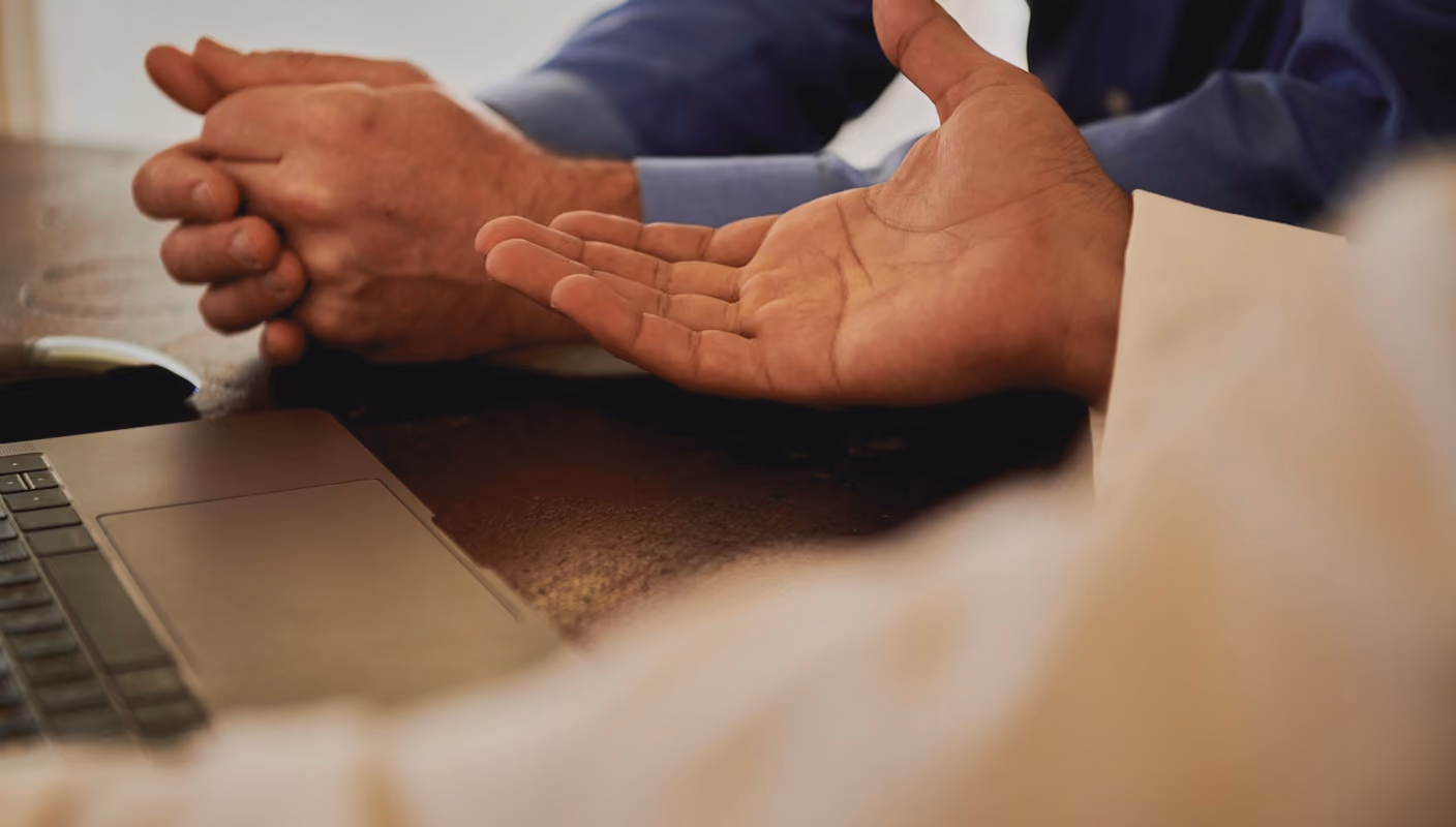 Two people sitting at a table with a laptop, one hand open and another clasped together.