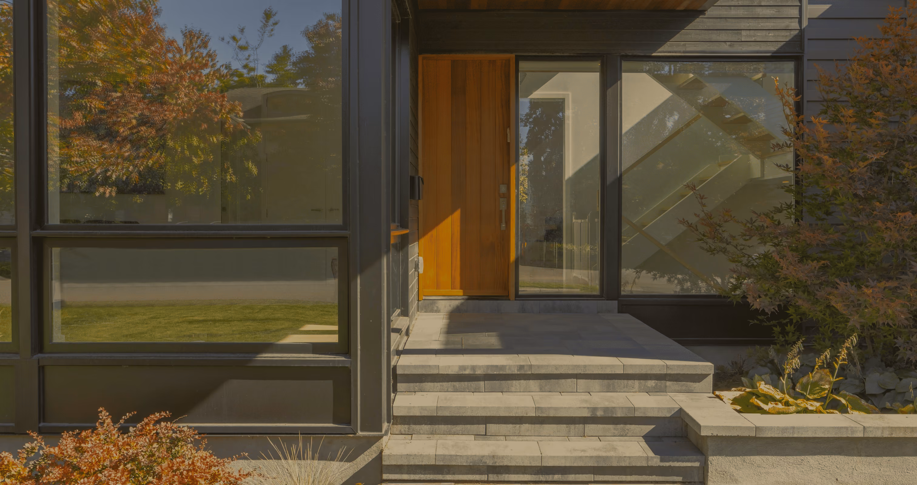 Modern house entrance with wooden door, large windows, stone steps, and surrounding autumn plants.