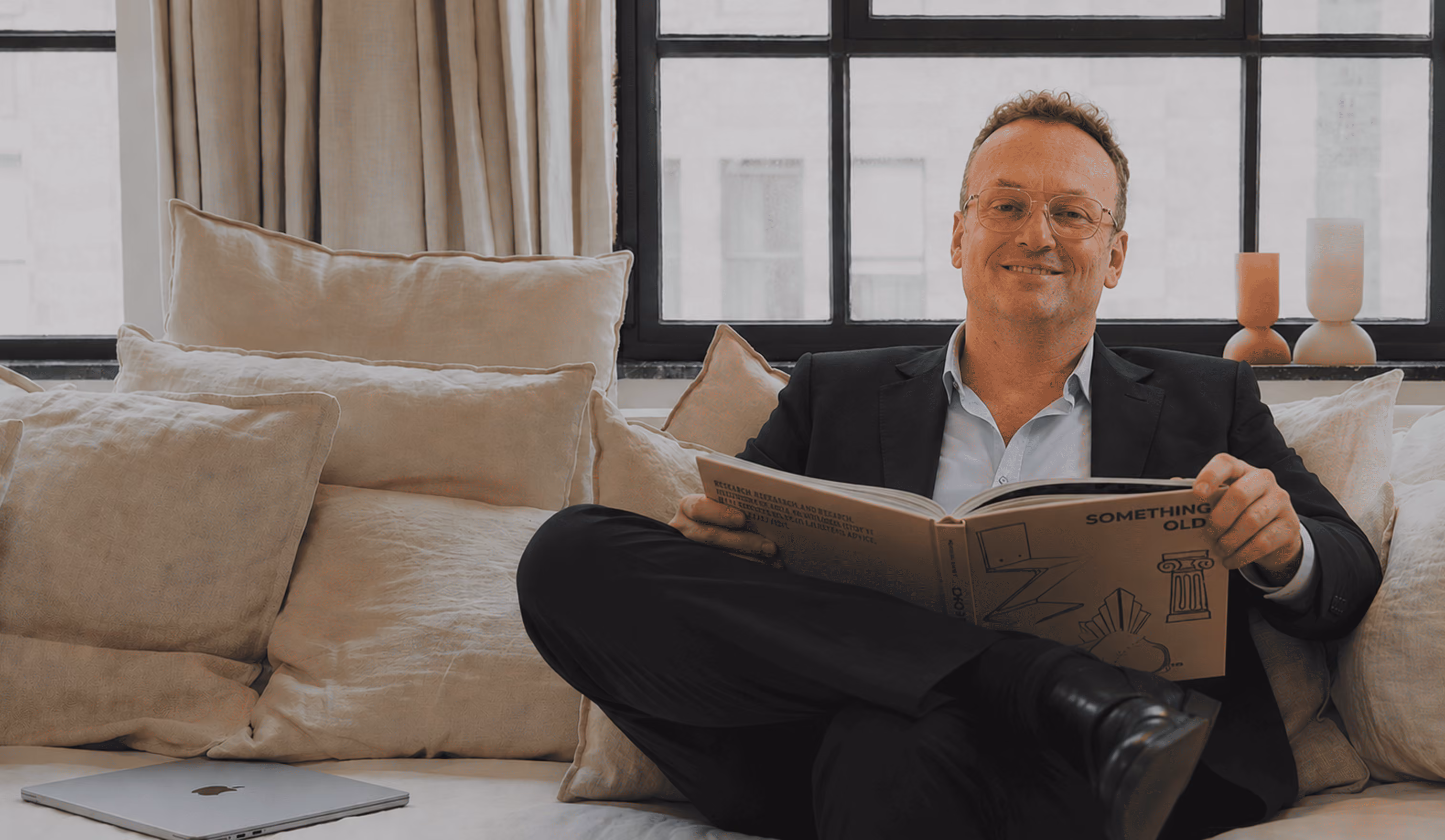 Smiling man in glasses and suit reading a book while sitting on a beige couch with cushions.