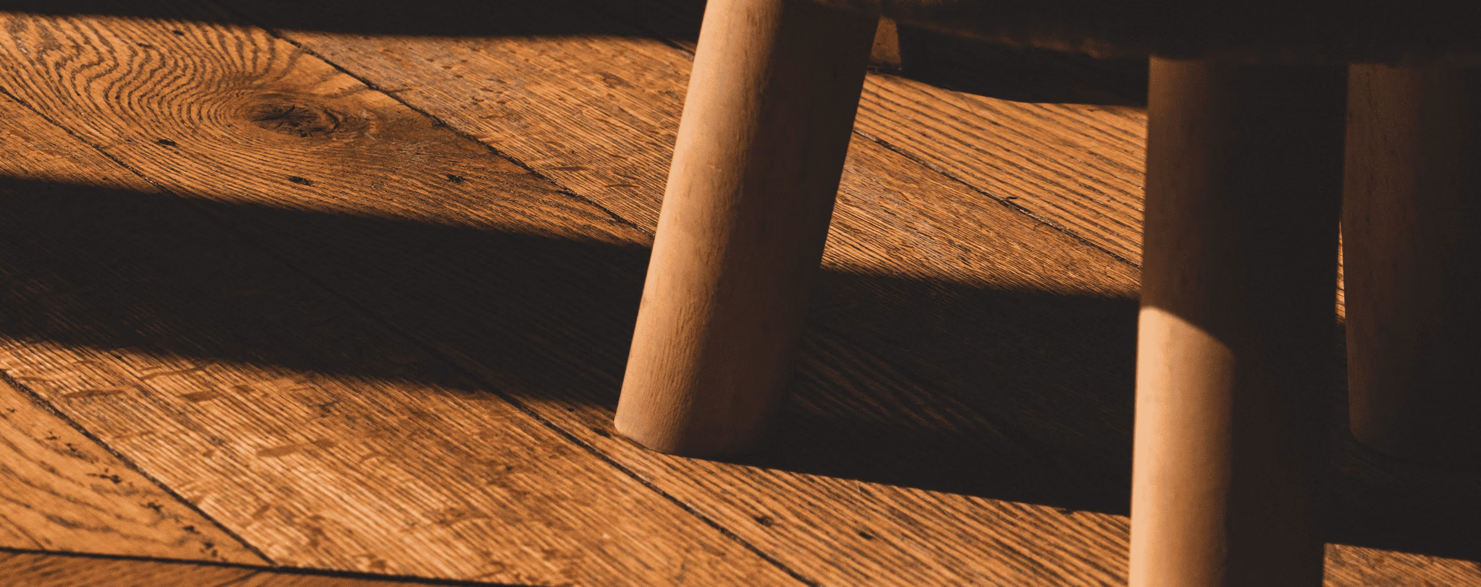 Close-up of wooden floorboards and two legs of a wooden chair casting shadows.