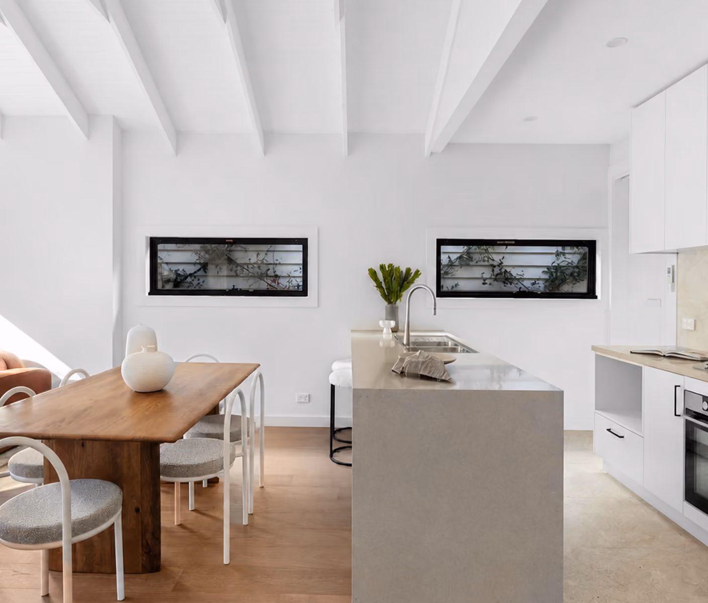Modern kitchen and dining area with wooden table, white chairs, sleek island, and horizontal windows.