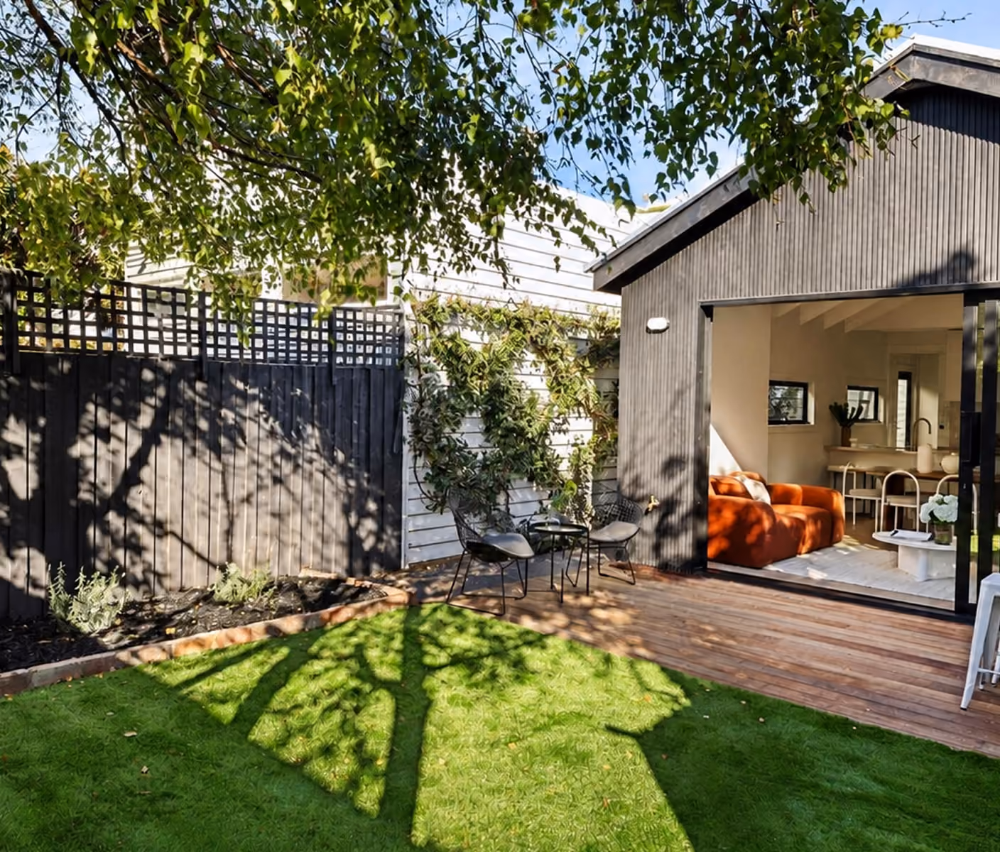 Backyard with green grass, wooden deck, black fence, two chairs and open house with orange sofa.
