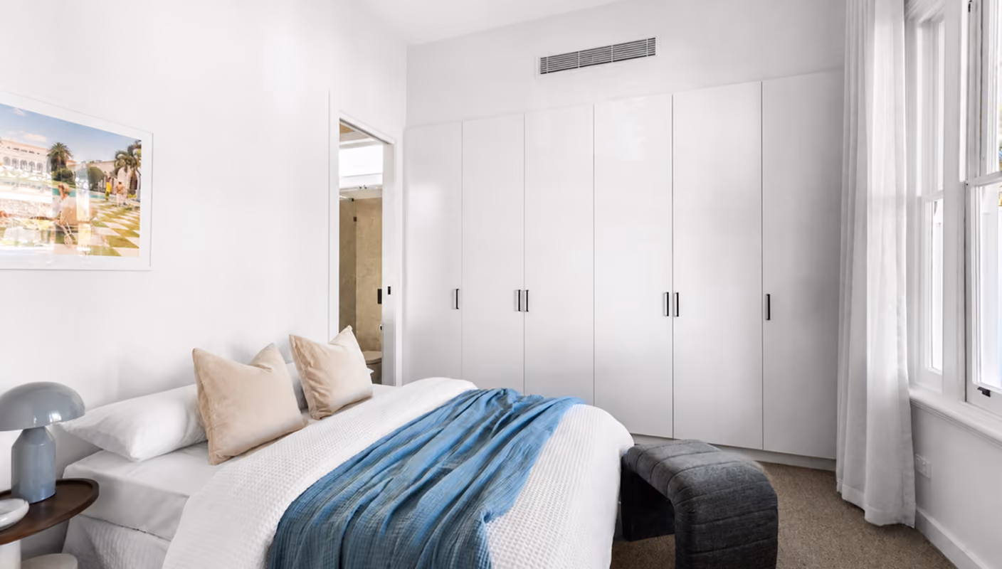 Bright bedroom with white walls, large wardrobe, double bed with beige pillows and blue throw.