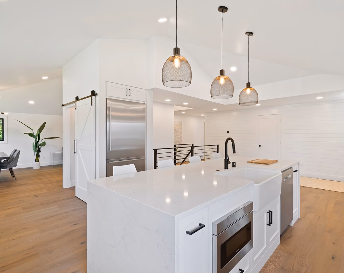 Modern white kitchen with large island, farmhouse sink, pendant lights, and stainless steel appliances.